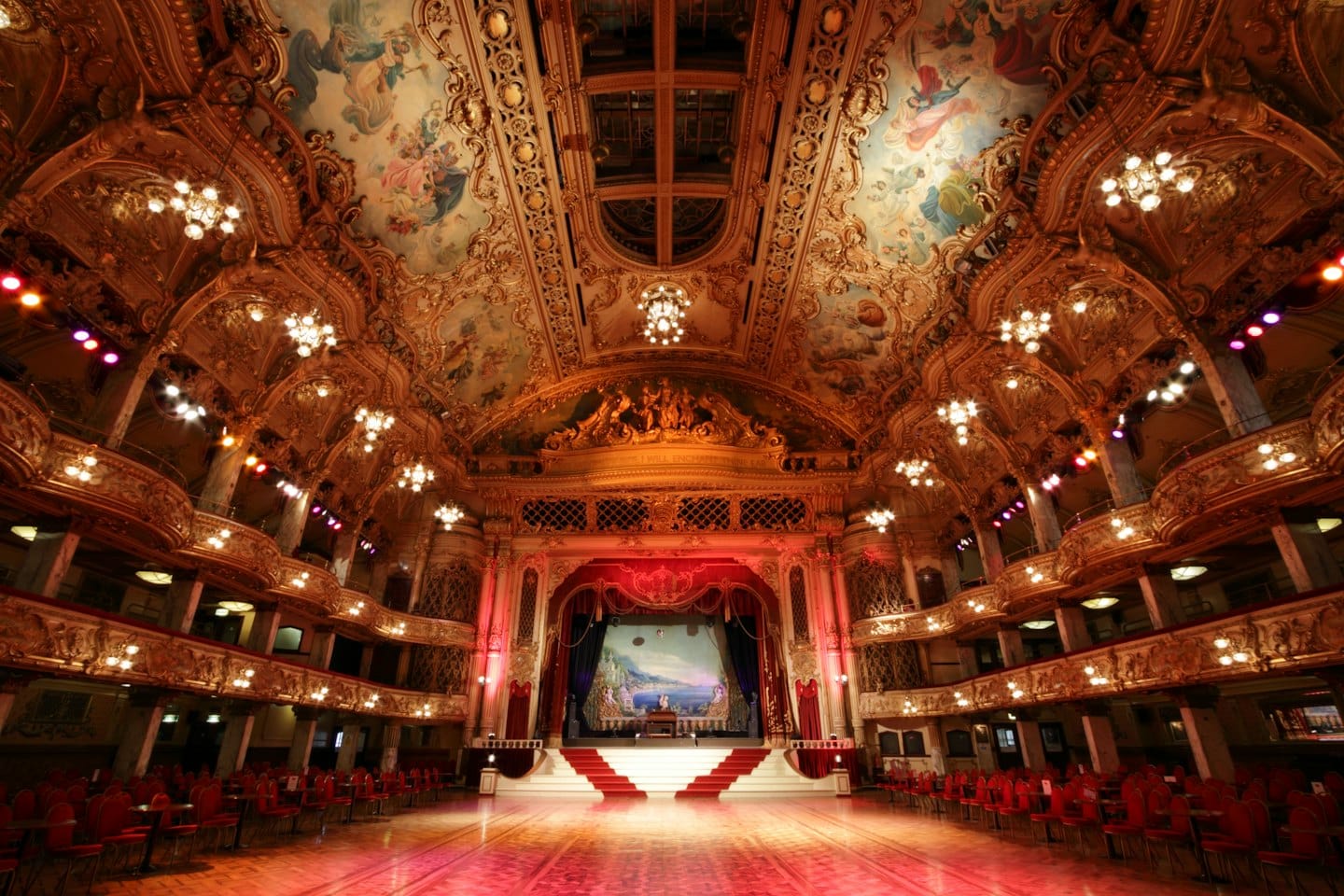Sala da ballo vuota e opulenta con balconi dorati e sedie rosse.